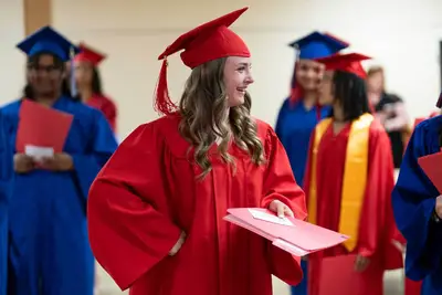 A young woman in a red graduation gown and cap smiles, holding a diploma folder.