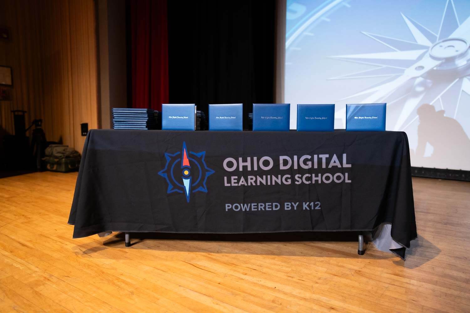 A table with a black cloth bearing the Ohio Digital Learning School logo is topped with neatly arranged blue diplomas