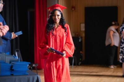 A graduate in a red cap and gown smiles, holding a diploma on stage.
