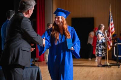 A young woman in a blue cap and gown smiles while shaking hands with a man on stage at a graduation ceremony.