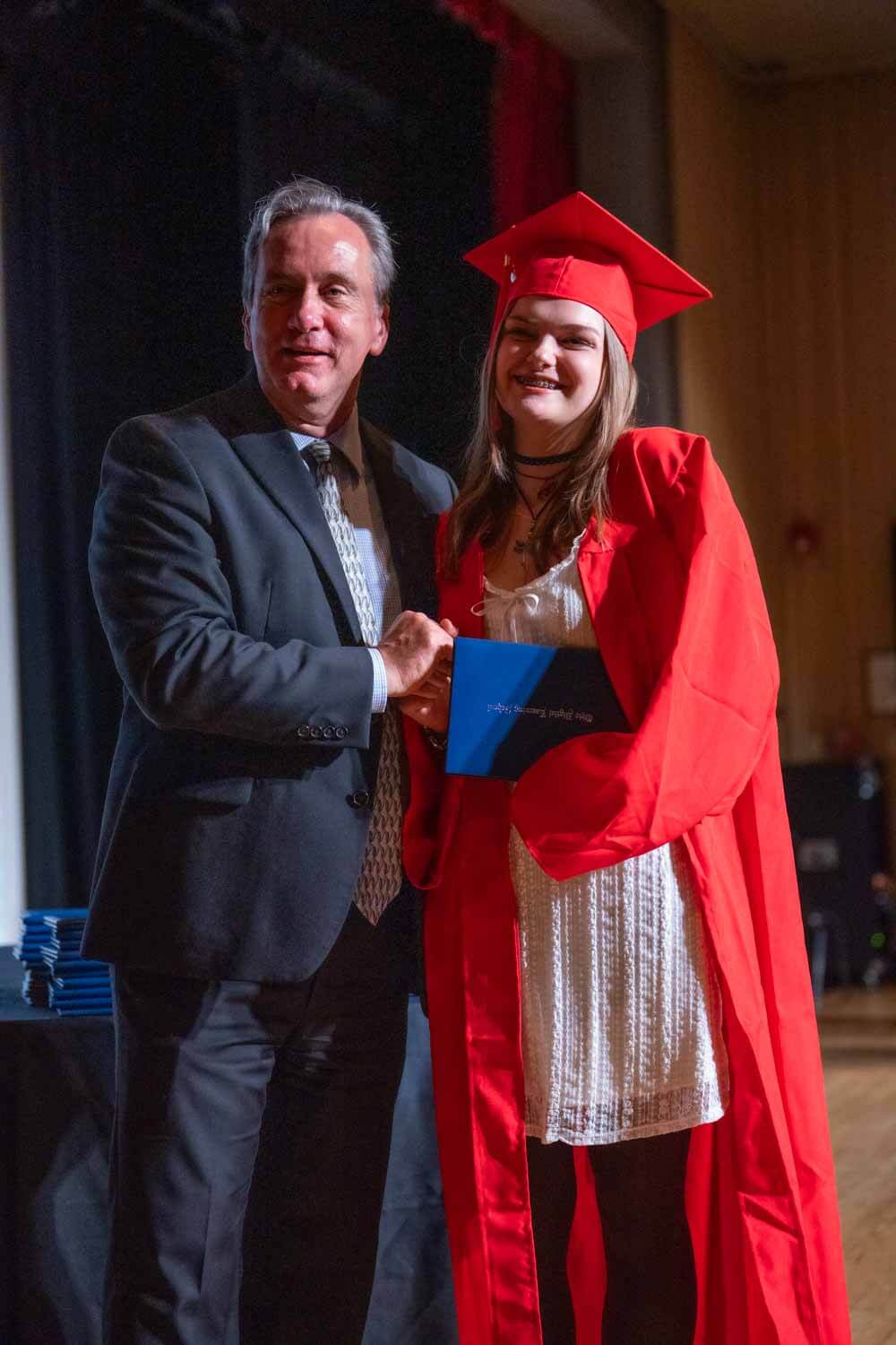 A graduate in a red cap and gown smiles as she shakes hands with a man in a suit on stage, holding a diploma.