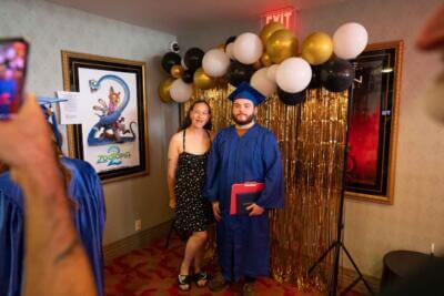 A graduate in a blue cap and gown poses with a woman in a black dress.