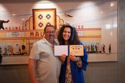 A smiling young woman in a blue graduation gown and cap holds a diploma, standing beside a man.