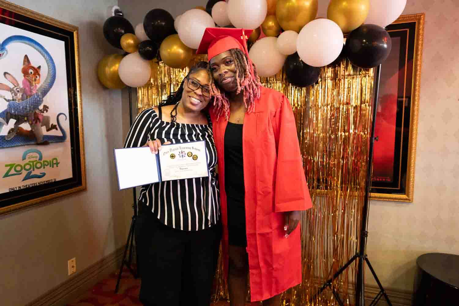 A graduate in a red cap and gown poses with a woman holding a certificate.