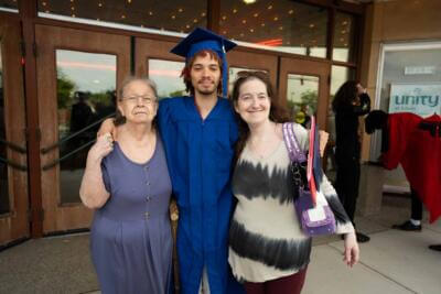 Graduate in a blue cap and gown stands smiling between an elderly woman and a middle-aged woman